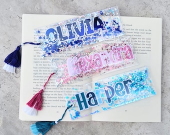 May include: Three rectangular clear acrylic bookmarks filled with colorful glitter and star-shaped sequins. Each bookmark features a custom name in bold lettering: OLIVIA in blue, alexandra in pink, and Harper in teal, each with a matching tassel.