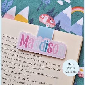 May include: A personalized bookmark with the name "Maddison" in pink and blue lettering. The bookmark is on a book page, with a light blue tab. The background features a camping theme with a camper, mountains, and a rainbow. "More Colors Available!" text is also visible.