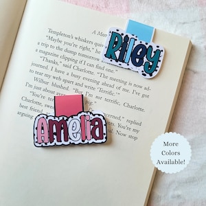May include: Two personalized name bookmarks, one with "Riley" in blue and white, the other with "Amelia" in pink and red, are placed on the pages of a book. The text "More Colors Available!" is also visible.
