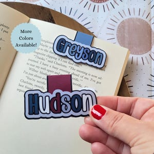 May include: Two personalized name bookmarks, one with the name "Greyson" in blue and white, and the other with "Hudson" in blue and red. The bookmarks are on a book page with the text "More Colors Available!"