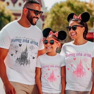May include: A family of three wearing white t-shirts with a graphic of Cinderella's castle and the text "Reach out and find your Happily Ever After". The shirts are in black and pink. The family is wearing Mickey Mouse ears.
