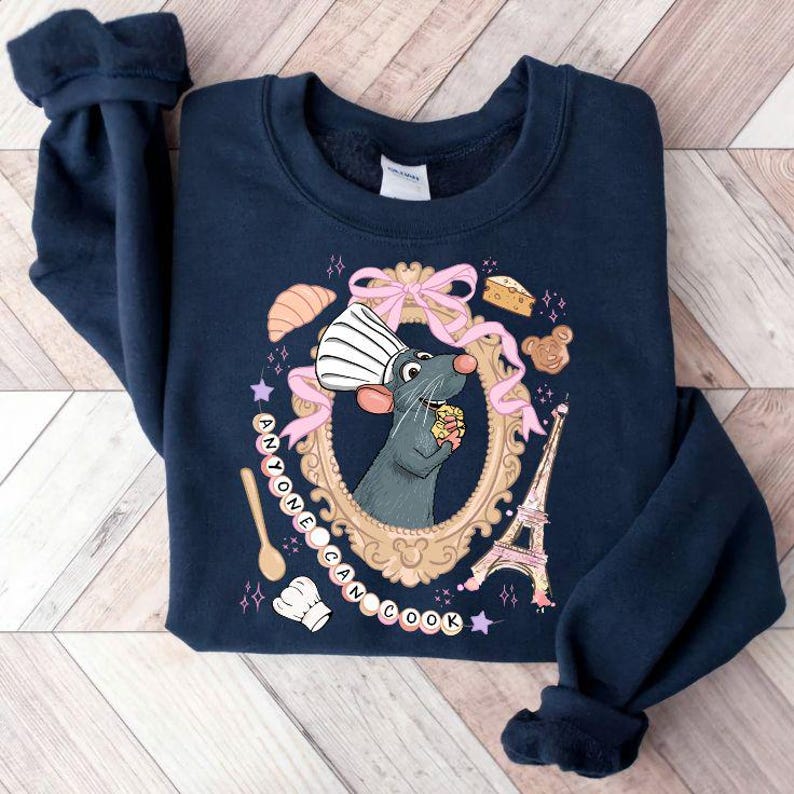 May include: Navy blue sweatshirt featuring a cartoon rat chef inside a gold frame with pink ribbons and pastries. The text "ANYONE CAN COOK" is written around the frame. Includes an Eiffel Tower illustration.