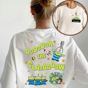 May include: White sweatshirt featuring a colorful graphic design from the movie Toy Story. The design includes the text "OooOooH THE CLAAAAAW" with cartoon aliens, a rocket, and a claw machine. The front of the sweatshirt has a small pocket design with the text "EST.1995".