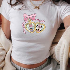 Op de afbeelding: Witte crop top met een cartoonafbeelding van Katrien Duck en Minnie Mouse in hartvormige medaillons. Het ontwerp bevat een roze strik met stippen en een gouden ketting. Een casual kledingstuk voor dagelijks gebruik.