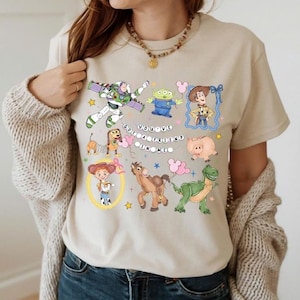 Puede incluir: Camiseta beige con personajes de dibujos animados coloridos de la película Toy Story. El diseño incluye a Buzz Lightyear, Woody, un extraterrestre y otros personajes, con el texto "You've Got a Friend in Me".