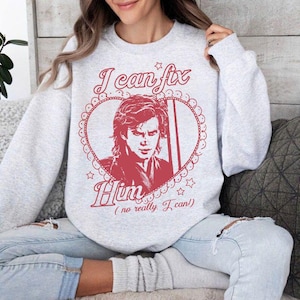 Op de afbeelding: Een lichtgrijs sweatshirt met een rode afbeelding. De afbeelding toont een portret in een hartvormige lijst, met de tekst "I can fix Him (no really, I can!)" in een scriptlettertype. Het sweatshirt heeft lange mouwen.