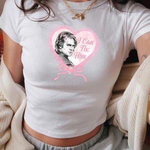 May include: White crop top with a pink heart graphic. The heart features a portrait and the text "I Can Fix Him". A pink bow is below the heart. The top is short-sleeved.