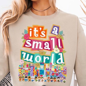 May include: Beige sweatshirt with the text "it's a small world" in colorful, block letters. The design includes floral accents and a whimsical illustration of a colorful cityscape with cartoon figures.