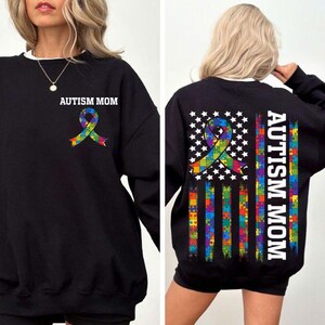 May include: Black sweatshirt with a white autism awareness ribbon and the text "Autism Mom" on the front. The back of the sweatshirt features a white American flag with puzzle pieces in the stripes and a white ribbon with puzzle pieces in the center. The text "Autism Mom" is printed vertically on the right side of the flag.