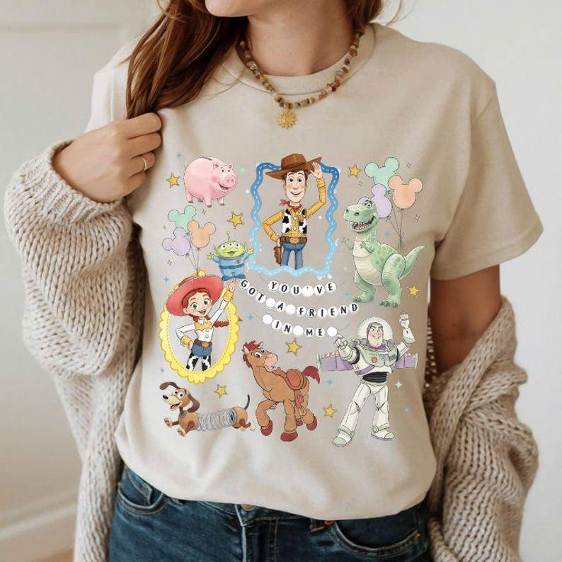 May include: Beige t-shirt featuring colorful cartoon characters from a popular animated movie. The design includes Woody, Buzz Lightyear, Rex, and other characters, with the text "YOU'VE GOT A FRIEND IN ME".