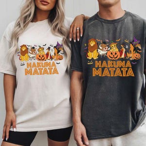 Puede incluir: Dos camisetas, una beige claro y otra gris antracita, con un diseño temático de Halloween. El diseño incluye personajes de dibujos animados, calabazas, murciélagos y el texto naranja "HAKUNA MATATA". Las camisetas son de talla grande.