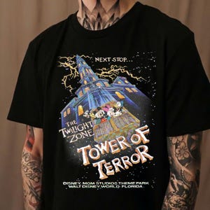 May include: Black t-shirt with a graphic of the Twilight Zone Tower of Terror ride at Disney's Hollywood Studios. The graphic features the ride's iconic tower, a lightning bolt, and the text "Next Stop... The Twilight Zone Tower of Terror".