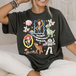 May include: Dark gray t-shirt featuring colorful cartoon characters from the Toy Story movie. The design includes Woody, Buzz Lightyear, Rex, Jessie, Hamm, and other characters. The shirt has a crew neck and short sleeves.