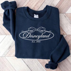 May include: A navy blue sweatshirt with the word "Disneyland" in white script inside an oval with a bow design. The text "Est. 1955" is below the oval. The sweatshirt is folded on a light brown wooden surface.