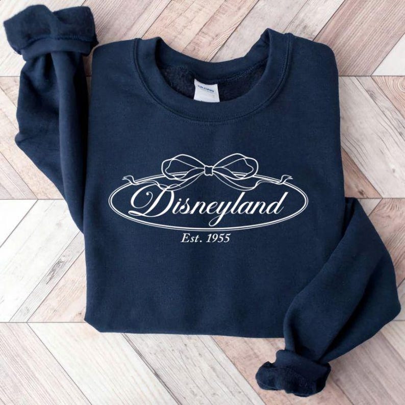 May include: A navy blue sweatshirt with the word "Disneyland" in white script inside an oval with a bow design. The text "Est. 1955" is below the oval. The sweatshirt is folded on a light brown wooden surface.