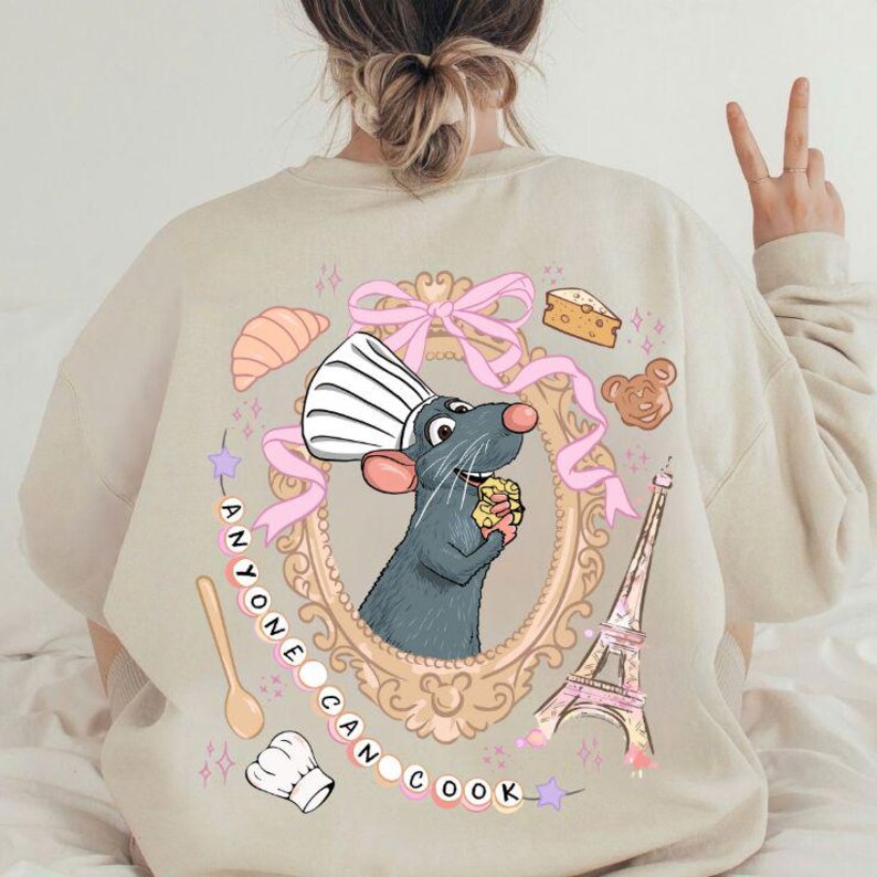 May include: Beige sweatshirt featuring a cartoon rat chef inside a gold frame, holding cheese. Pink ribbon, Eiffel Tower, and text "ANYONE CAN COOK" surround the frame. Additional illustrations include a croissant, cheese, and a bear-shaped pastry.