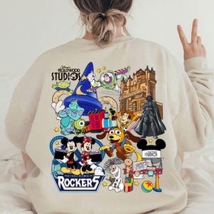 May include: Beige sweatshirt featuring a colorful graphic of Disney's Hollywood Studios characters, including Mickey and Minnie Mouse, Buzz Lightyear, and Darth Vader. The design includes the text "Hollywood Studios" and "Rockers".