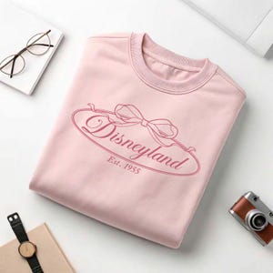 May include: A light pink sweatshirt with the word "Disneyland" in a cursive font, framed by a bow design. The text "Est. 1955" is below the word Disneyland. The sweatshirt is folded neatly on a white surface.