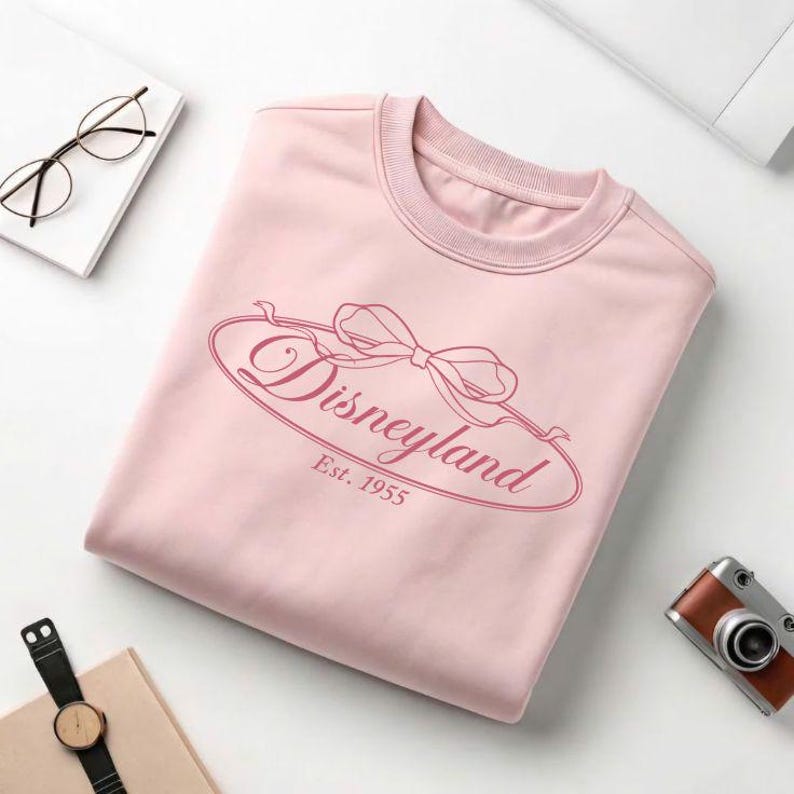 May include: A light pink sweatshirt with the word "Disneyland" in a cursive font, framed by a bow design. The text "Est. 1955" is below the word Disneyland. The sweatshirt is folded neatly on a white surface.