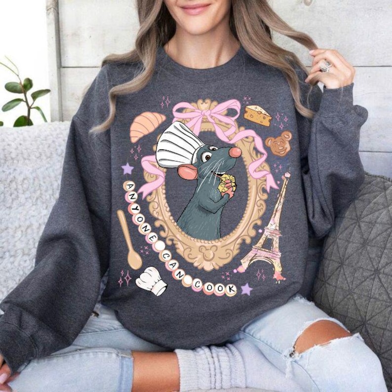 May include: Dark gray sweatshirt featuring a cartoon rat chef inside a gold frame with a pink bow. The design includes a spoon, chef's hat, Eiffel Tower, and the text "ANYONE CAN COOK". The sweatshirt is a cozy, casual style.