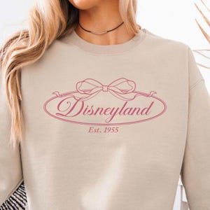 May include: Beige sweatshirt with a pink Disneyland logo. The logo features a bow and the word "Disneyland" in a cursive font, with "Est. 1955" below. A comfortable, casual top for Disney fans.