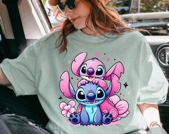 Könnte beinhalten: Ein hellblaues T-Shirt mit einem Motiv von Stitch und Angel aus dem Disney-Film Lilo & Stitch. Stitch ist blau und Angel ist rosa. Beide lächeln und haben große Augen. Das Shirt hat einen hellgrünen Hintergrund.