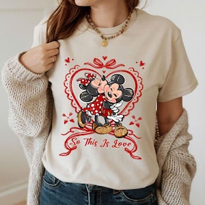 Puede incluir: Camiseta beige con un gráfico de Mickey y Minnie Mouse abrazándose dentro de un corazón rojo. Minnie lleva un vestido y un lazo de lunares rojos. El texto "So This Is Love" está debajo. Prenda de vestir informal.