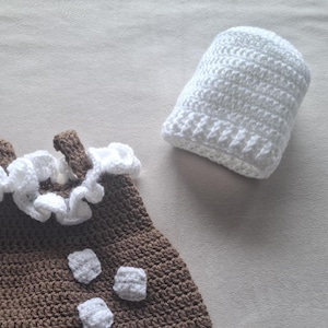 May include: A brown crocheted dress featuring a white ruffled neckline and three small white crocheted squares on the front, accompanied by a matching white crocheted hat. The set is crafted with a visible yarn texture throughout each piece.
