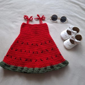 May include: A red crochet dress with a dark green ruffled hem and black stitched details resembling watermelon seeds, featuring two tie-top shoulder straps, accompanied by a pair of white shoes with bows and round white-framed sunglasses.