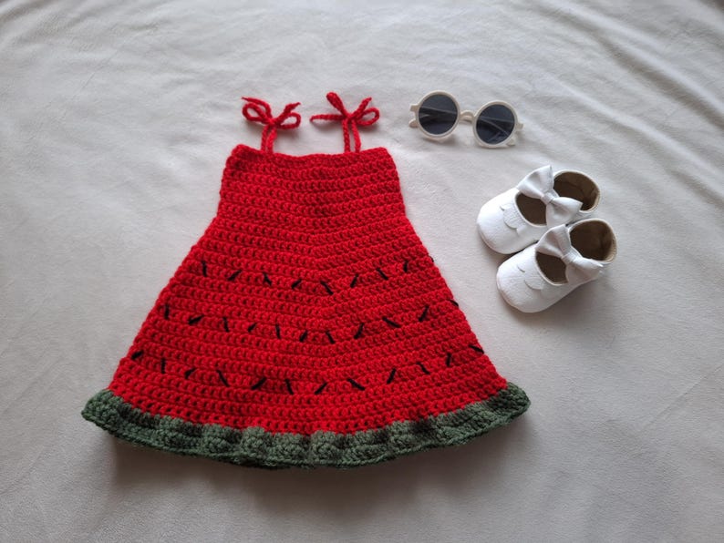 May include: A red crochet dress with a dark green ruffled hem and black stitched details resembling watermelon seeds, featuring two tie-top shoulder straps, accompanied by a pair of white shoes with bows and round white-framed sunglasses.