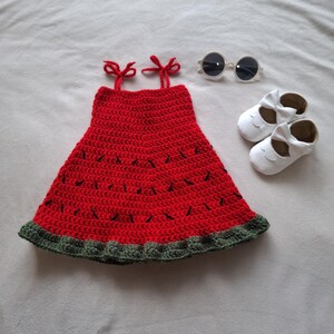 May include: A red crochet dress with black stitched details, a dark green ruffled hem, and two shoulder straps tied in bows. The set includes a pair of white round-frame sunglasses and two white leather-like shoes with decorative bows on the front.