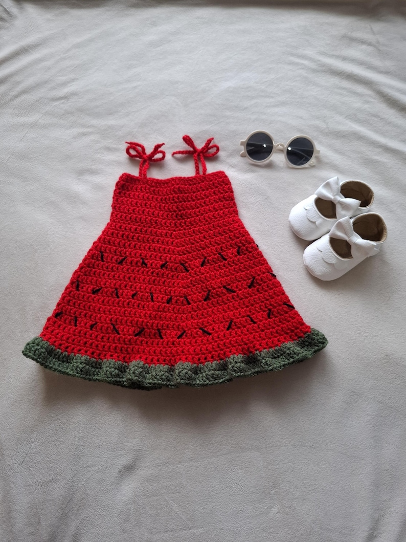 May include: A red crochet dress with black stitched details, a dark green ruffled hem, and two shoulder straps tied in bows. The set includes a pair of white round-frame sunglasses and two white leather-like shoes with decorative bows on the front.
