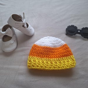 May include: A crochet beanie featuring horizontal stripes of white, orange, and yellow, resembling candy corn. Accompanying the hat are a pair of white shoes with bow accents and a pair of black sunglasses with round, scalloped-edge frames.