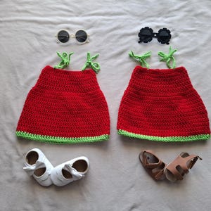 May include: Two red crochet dresses with green trim and straps, paired with white shoes and brown sandals. Two pairs of sunglasses, one white and one black, are also included. The items are arranged on a light gray surface.