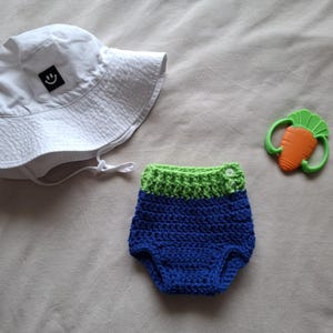 May include: A white baby sun hat with a black smiley face patch, a blue and green crochet diaper cover, and a carrot-shaped teether. The items are arranged on a neutral-colored surface.