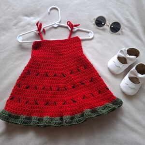 May include: A red crochet dress with black stitched details resembling watermelon seeds and a dark green ruffled hem, displayed on a white hanger, accompanied by a pair of white baby shoes with bows and round white-framed sunglasses.