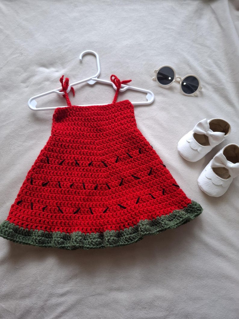 May include: A red crochet dress with black stitched details resembling watermelon seeds and a dark green ruffled hem, displayed on a white hanger, accompanied by a pair of white baby shoes with bows and round white-framed sunglasses.