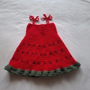 May include: A sleeveless crochet dress featuring a vibrant red bodice with small black stitched accents resembling seeds. The garment has two thin red shoulder straps tied in bows and a wide, textured dark green border along the bottom hem.