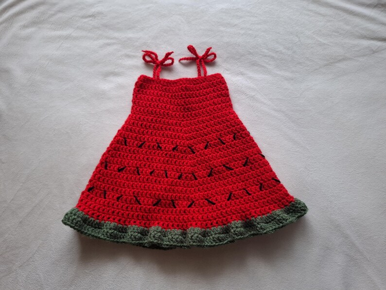 May include: A sleeveless crochet dress featuring a vibrant red bodice with small black stitched accents resembling seeds. The garment has two thin red shoulder straps tied in bows and a wide, textured dark green border along the bottom hem.