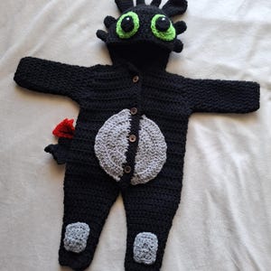 May include: A black crochet baby outfit resembling a dragon, featuring a hood with green eyes and ear-like protrusions. The outfit includes a gray belly patch, button closures, and a red tail fin. Gray patches are on the knees.