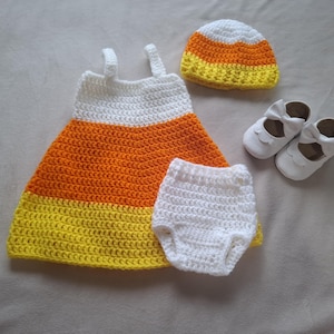 Puede incluir: Un conjunto de ganchillo que incluye un vestido sin mangas, un gorro y unos bombachos con un esquema de colores blanco, naranja y amarillo. El conjunto incluye un par de zapatos blancos con un detalle de lazo en la parte delantera de hilo texturizado.