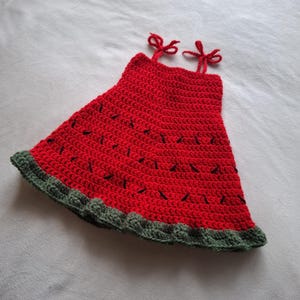 May include: A sleeveless crochet dress featuring a vibrant red bodice with small black stitched accents resembling watermelon seeds. The hem is finished with a textured dark green border, and the top includes two thin red straps tied into bows.