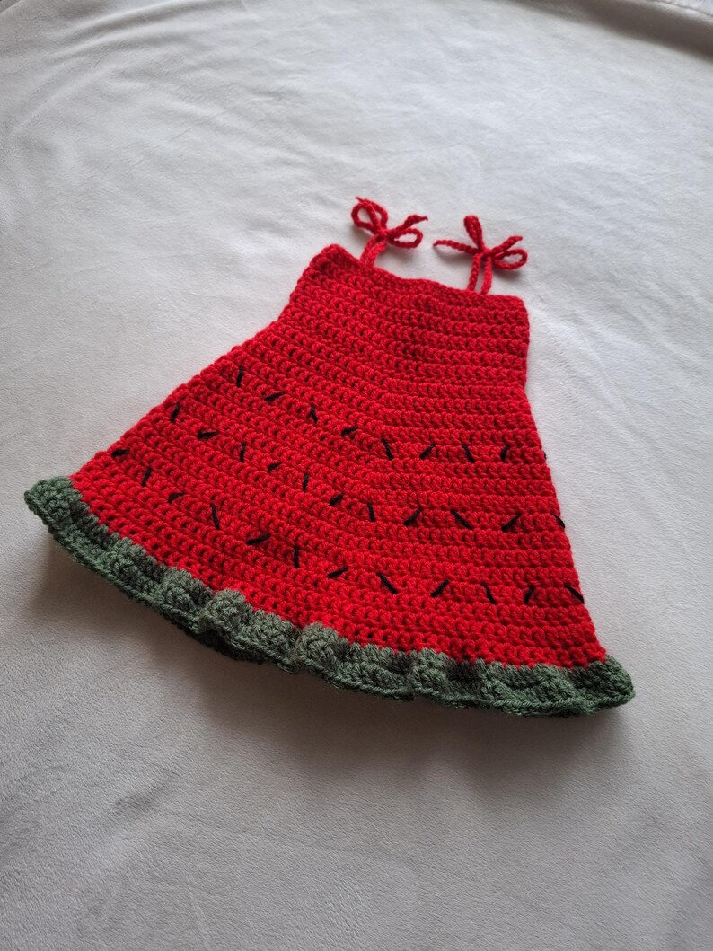 May include: A sleeveless crochet dress featuring a vibrant red bodice with small black stitched accents resembling watermelon seeds. The hem is finished with a textured dark green border, and the top includes two thin red straps tied into bows.