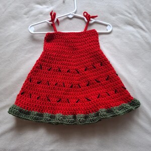 May include: A sleeveless crochet dress featuring a vibrant red bodice with scattered black yarn stitches resembling watermelon seeds. The hem is finished with a textured dark green border, and the top includes two thin red straps tied at the shoulders.