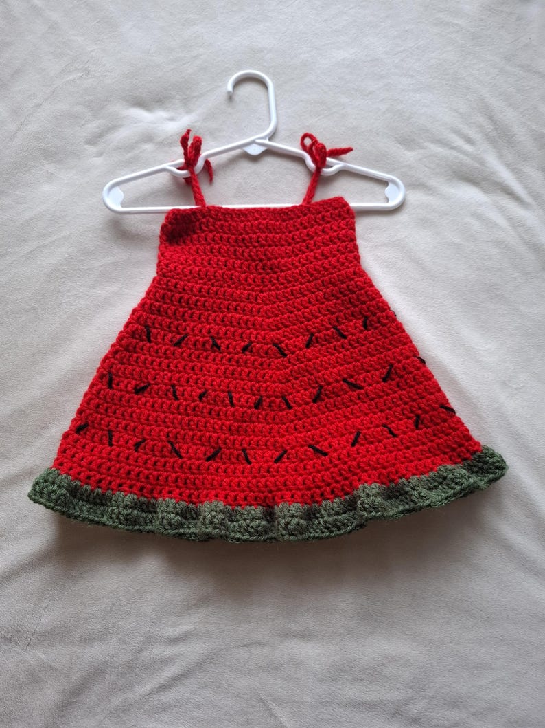 May include: A sleeveless crochet dress featuring a vibrant red bodice with scattered black yarn stitches resembling watermelon seeds. The hem is finished with a textured dark green border, and the top includes two thin red straps tied at the shoulders.