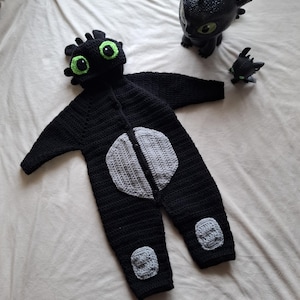 Puede incluir: Un conjunto de bebé de ganchillo negro que se asemeja a un dragón, con una capucha con ojos verdes y una mancha en el vientre gris. El atuendo incluye parches en las rodillas a juego. También hay una figura de dragón negro y una figura de dragón más pequeña.