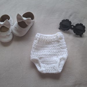 May include: A pair of white shoes with scalloped edges and bows, a white crocheted diaper cover with a small button detail, and a pair of black sunglasses with round, flower-shaped frames arranged on a neutral surface.