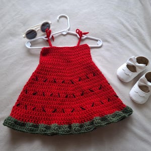 May include: A red crochet dress with black stitched details resembling watermelon seeds and a dark green ruffled hem, displayed on a white plastic hanger alongside a pair of white round-frame sunglasses and white leather-style baby shoes.