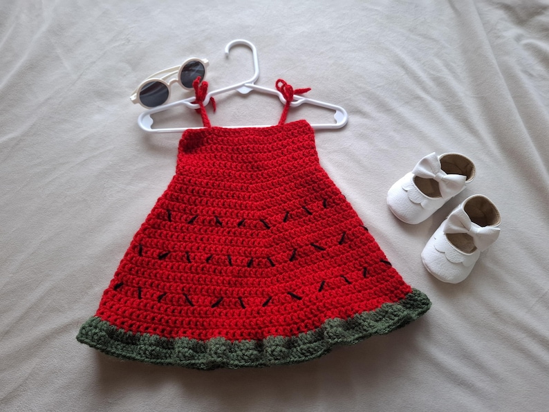 May include: A red crochet dress with black stitched details resembling watermelon seeds and a dark green ruffled hem, displayed on a white plastic hanger alongside a pair of white round-frame sunglasses and white leather-style baby shoes.