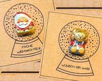 May include: Two brown cards with snow globe illustrations and chocolate figures inside. One card features a Santa Claus figure, the other a teddy bear. The text "FROHE WEIHNACHTEN!" and "WÜNSCH DIR WAS!" are printed below the globes.